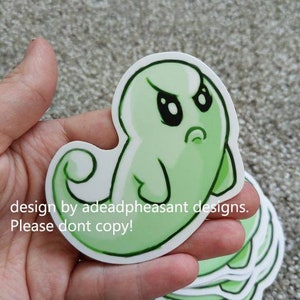 May include: Green ghost sticker with an angry expression. The sticker is on a hand and there are more stickers stacked behind it.