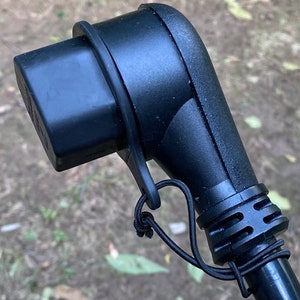 May include: Black electrical plug with a black rubber strap for securing the cord. The plug has a black rubber cover over the prongs.
