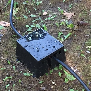 May include: A black electrical outlet box with a black cord attached. The box is sitting on the ground and is wet from rain.