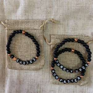 May include: Two black beaded bracelets with gold accents. The first bracelet says "I love Eagles" with a small heart charm. The second bracelet has three bands that say "Hungry Dogs", "Run" and "Faster".