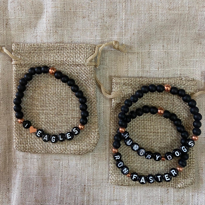 May include: Two black beaded bracelets with gold accents. The first bracelet says "I love Eagles" with a small heart charm. The second bracelet has three bands that say "Hungry Dogs", "Run" and "Faster".