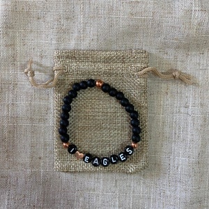 May include: Black beaded bracelet with gold accents and white letter beads spelling "I love Eagles". The bracelet is in a small burlap bag.