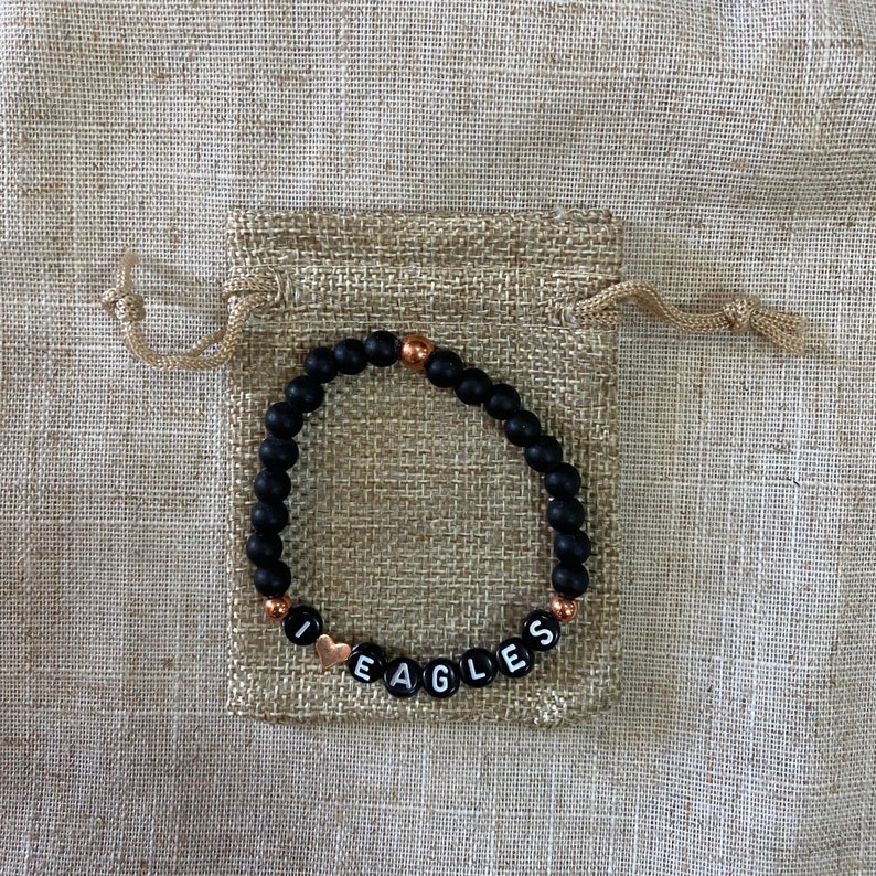 May include: Black beaded bracelet with gold accents and white letter beads spelling "I love Eagles". The bracelet is in a small burlap bag.