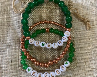 May include: Set of four bracelets with green and copper beads. The bracelets feature white beads with gold lettering spelling "GO BIRDS" and "EAGLES".