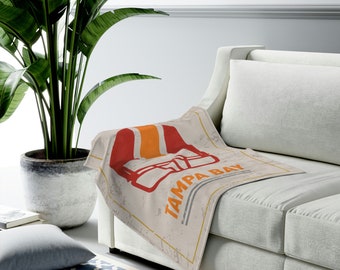 May include: A beige throw blanket with a distressed design featuring a red and orange football helmet and the words "TAMPA BAY" in orange. The blanket is draped over the arm of a white sofa.