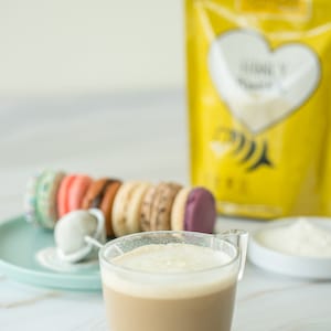 May include: A glass of coffee with a layer of foam on top, next to a plate of colorful macarons and a bag of gluten-free baking mix.