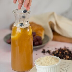 May include: A glass bottle filled with a light brown liquid, possibly tea or juice, with blueberries floating in it. The bottle is on a white surface with a book and a bowl of white powder.