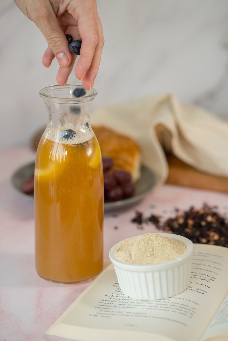 May include: A glass bottle filled with a light brown liquid, possibly tea or juice, with blueberries floating in it. The bottle is on a white surface with a book and a bowl of white powder.