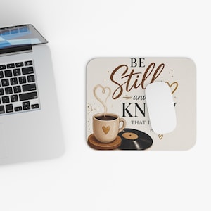 Può includere: Un tappetino per mouse rettangolare con sfondo beige e il testo BE STILL AND KNOW THAT I AM. Il design include una tazza di caffè con un cuore, vapore a forma di cuore, un disco in vinile e piccole icone a forma di cuore in marrone e nero.