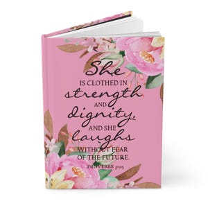 May include: Pink journal with floral accents and the text "She is clothed in strength and dignity, and she laughs without fear of the future. Proverbs 31:25."