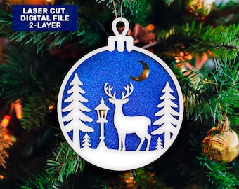 Può includere: Un ornamento natalizio tagliato al laser a due strati con una scena invernale. L'ornamento presenta un cervo bianco, alberi e un lampione su uno sfondo blu glitterato. Il testo "LASER CUT DIGITAL FILE 2-LAYER" è in alto a sinistra.
