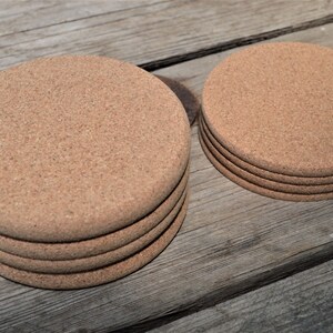 May include: A set of five round cork coasters, with a smaller set of four coasters in the background. The coasters are a light brown color and have a natural cork texture.