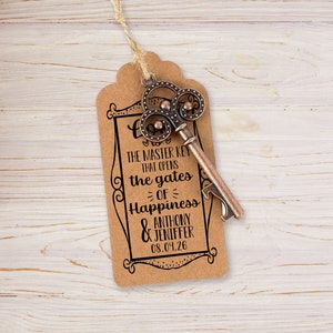 Puede incluir: Una etiqueta de papel marr&oacute;n con un marco negro decorativo y el texto "This is the master key that opens the gates of happiness Anthony & Jennifer 08.04.26". Un abridor de botellas vintage en forma de llave est&aacute; unido a la etiqueta.