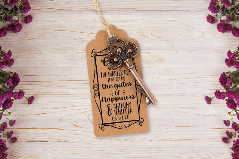 Puede incluir: Una etiqueta de papel marr&oacute;n con un marco negro decorativo y el texto "This is the master key that opens the gates of happiness Anthony & Jennifer 08.04.26". Un abridor de botellas vintage en forma de llave est&aacute; unido a la etiqueta.