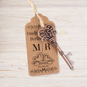 Puede incluir: Una etiqueta de papel marr&oacute;n con el texto "Finding Perfect" y "M|R" con dos corazones y el texto "Married 01.06.28" impreso en ella. Un abridor de botellas con forma de llave vintage est&aacute; unido a la etiqueta con un cordel.