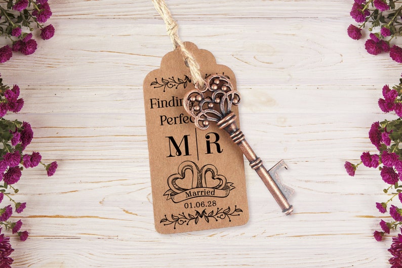 Puede incluir: Una etiqueta de papel marr&oacute;n con el texto "Finding Perfect" y "M|R" con dos corazones y el texto "Married 01.06.28" impreso en ella. Un abridor de botellas con forma de llave vintage est&aacute; unido a la etiqueta con un cordel.