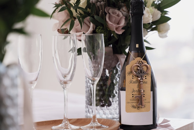 Puede incluir: Una botella de champ&aacute;n con un abridor de botellas de llave dorada adjunto. La etiqueta en el abridor de botellas dice "Katherine & Joshua" y "Gracias por celebrar con nosotros".