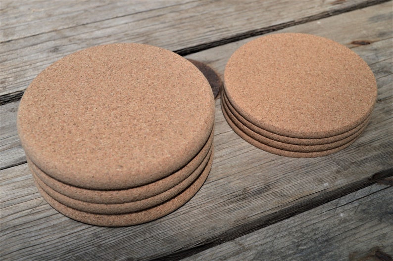 May include: A set of five round cork coasters, with a smaller set of four coasters in the background. The coasters are a light brown color and have a natural cork texture.