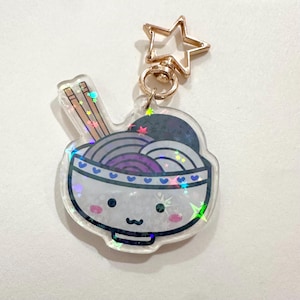 May include: A clear acrylic keyring featuring a cartoon ramen bowl with a smiling face, chopsticks, and noodles in shades of purple and grey. The keyring has a gold star-shaped clasp and a gold ring. The bowl is decorated with blue hearts.