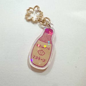 May include: A clear acrylic keychain with a Kewpie mayonnaise bottle design. The bottle is beige with pink accents and Japanese text. The keychain has a gold-coloured flower-shaped clasp.