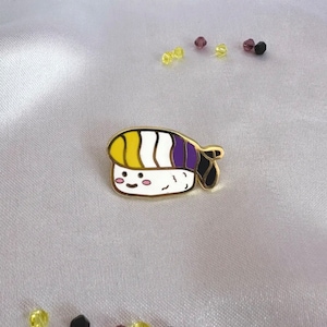 Puede incluir: Un pin de esmalte de color dorado con forma de rollo de sushi con un diseño amarillo, blanco y morado. El pin tiene una cara sonriente y está sobre un fondo de satén blanco.
