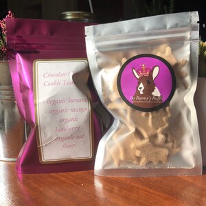May include: A purple resealable bag with a label that reads "Chocolate Chip Cookie Tiaras" and lists the ingredients: organic banana, organic mango, organic blueberry, and organic oat flour.  The bag is next to a clear resealable bag filled with small, star-shaped, light brown treats. The clear bag has a round sticker with a cartoon bunny wearing a crown and the text "Bo Bunny's Buffet Everybun needs a crown!"