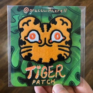 以下が含まれることがあります： オレンジ色の目をした黄色と黒の刺繍のアイロン接着パッチ。パッチは緑色の背景にオレンジ色と黒色の「TIGER PATCH」の文字が書かれている。
