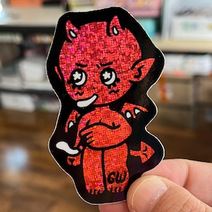 Peut inclure: Autocollant rouge représentant un diable avec une finition pailletée. Le diable de dessin animé a un visage souriant, des cornes et des ailes, soulignés en noir. L'autocollant porte les initiales "GW" en bas.