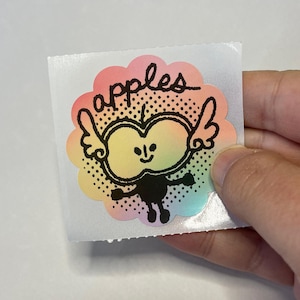 May include: A colorful sticker featuring a cartoon apple character with wings and the word "apples" in script. The apple has a smiling face and is outlined in black. The sticker has a scalloped edge with a rainbow gradient.