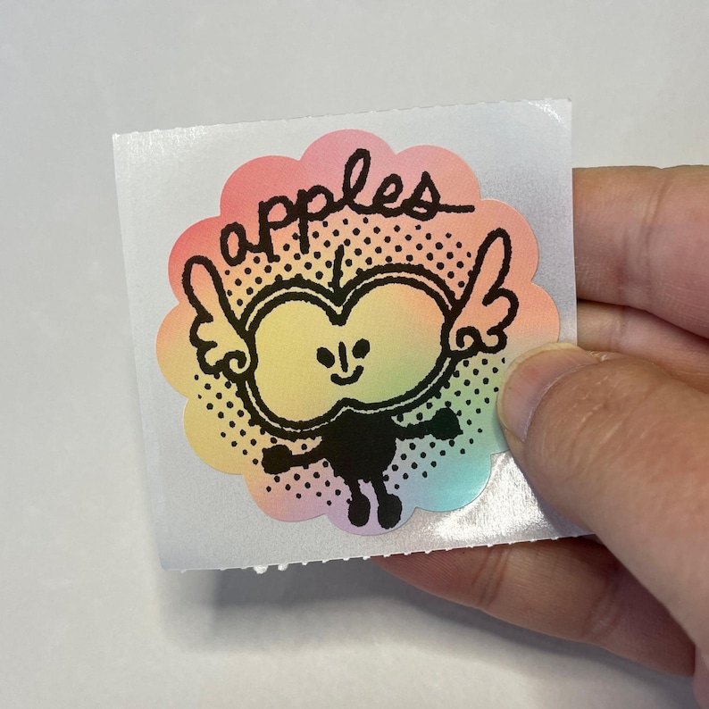 May include: A colorful sticker featuring a cartoon apple character with wings and the word "apples" in script. The apple has a smiling face and is outlined in black. The sticker has a scalloped edge with a rainbow gradient.