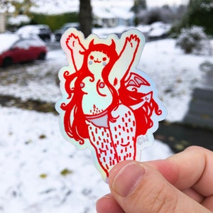 Puede incluir: Una pegatina holográfica roja y blanca de una mujer de dibujos animados con cuernos, alas y cola. La pegatina está sobre un fondo nevado.