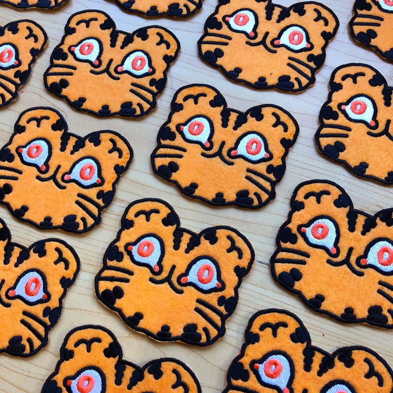 May include: A collection of orange and black felt tiger face patches with white eyes and orange pupils. The patches are arranged in a staggered pattern on a light brown wooden surface.