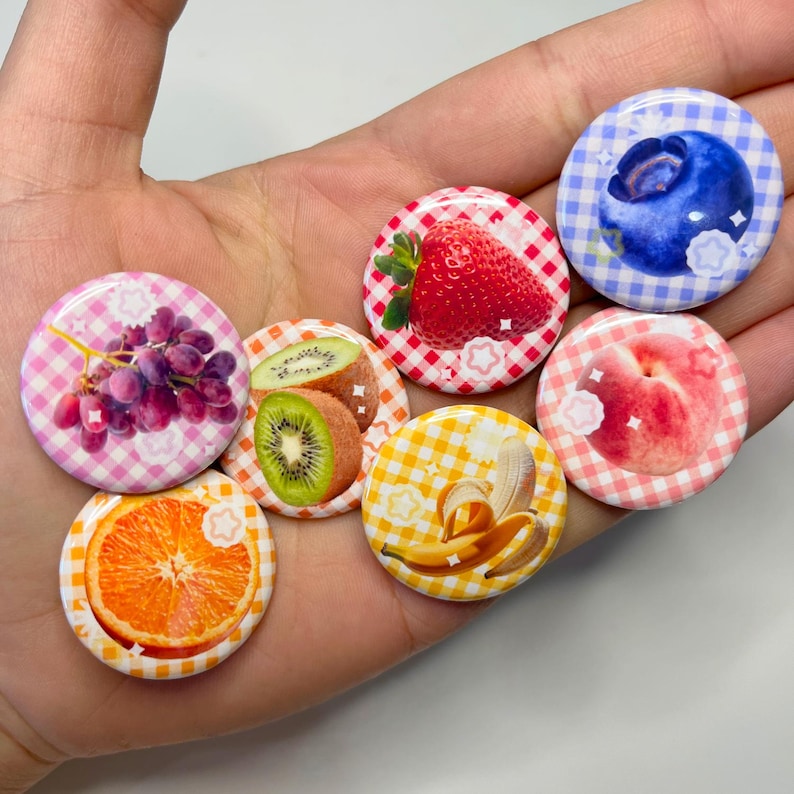 May include: A collection of round, colorful buttons featuring fruit designs. The buttons showcase images of grapes, kiwi, strawberries, oranges, bananas, peaches, and blueberries, set against gingham patterns. Each button has a star-shaped accent.