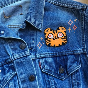May include: A blue denim jacket with a patch of an orange tiger face with black stripes and red eyes. The patch is surrounded by small white and pink stars.