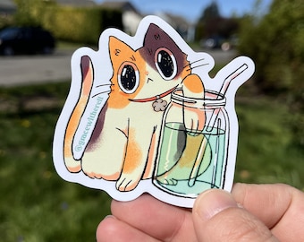 Può includere: Un gatto calico con un collare verde e un ciondolo sta tenendo un barattolo di vetro con un liquido verde e una cannuccia. Il gatto sta guardando la telecamera con un'espressione felice. Lo sfondo è sfocato e mostra un cielo blu e erba verde.