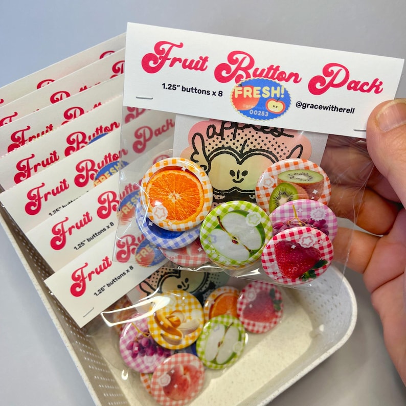 May include: A pack of fruit-themed buttons, each 1.25 inches in diameter, featuring images of oranges, apples, strawberries, and other fruits. The packaging includes the text "Fruit Button Pack" and "FRESH!" in a playful font. The buttons are displayed in a white tray.