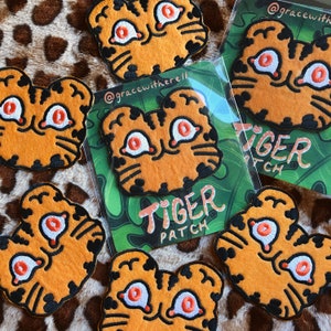 May include: A set of seven orange and black embroidered iron-on patches featuring a cartoon tiger with large eyes. The patches are packaged in clear plastic bags with a green and white background featuring the text "Tiger Patch".