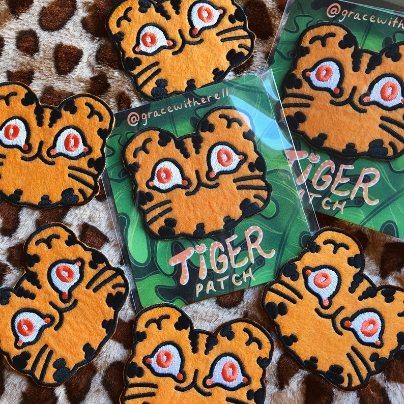 May include: A set of seven orange and black embroidered iron-on patches featuring a cartoon tiger with large eyes. The patches are packaged in clear plastic bags with a green and white background featuring the text "Tiger Patch".