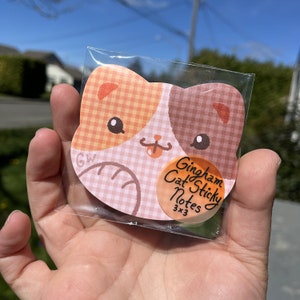 May include: A package of gingham cat sticky notes. The notes are shaped like a cat's head with a pink, orange, and brown gingham pattern. The package has a sticker on it that says "Gingham Cat Sticky Notes 3x3".