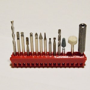 May include: A red plastic holder with 18 slots for holding small drill bits and sanding attachments. The holder is filled with a variety of bits, including a drill bit, a sanding drum, and several different types of grinding bits.