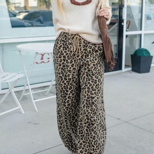 May include: A woman wearing a cream-colored sweater with a brown horseshoe design and leopard print pants. The pants have a drawstring waist and a wide leg. She is also wearing a brown shoulder bag and tan shoes.