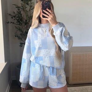 May include: A light blue and white long-sleeve sweatshirt and matching shorts set featuring a patchwork design with various patterns including floral, gingham, and polka dots. The sweatshirt has a crew neckline and ribbed cuffs, while the shorts feature an elasticized waistband with a drawstring.