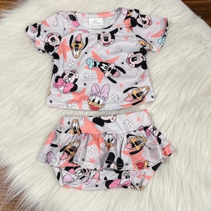 May include: A two-piece baby outfit featuring a short-sleeved top and ruffled shorts. The set is white with a repeating pattern of Mickey Mouse, Minnie Mouse, Donald Duck, and Daisy Duck, along with pink stars and black stars.