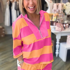 May include: A pink and orange striped two-piece outfit. The top is a hooded pullover with a quarter-zip closure. The shorts have an elastic waistband. The outfit is made of a soft, lightweight material.