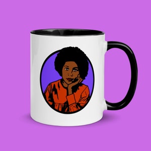 May include: White ceramic mug with a black interior and handle. The mug features a pop art style illustration of a person with dark skin, an afro, and an orange dress, set against a purple circle.