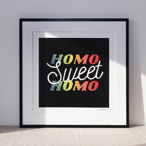 homo sweet homo linocut print in a black frame. the homo lettering is rainbow color while the SWEET is white allwitha black backdrop