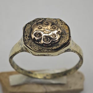 May include: A gold-toned ring featuring a textured, dark, octagonal face. Centered on the face is a raised, polished gold-coloured circular element with six small, recessed indentations arranged in a floral-like pattern.