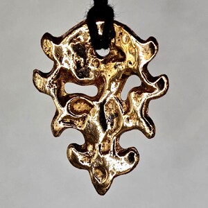 May include: A gold-colored pendant with an intricate, organic design. The pendant is suspended from a black cord. The design features a series of interconnected shapes and open spaces, creating a textured appearance. The pendant has a vintage or antique aesthetic.