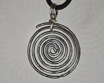 May include: A circular silver-toned metal pendant featuring a continuous spiral design. The pendant is suspended from a black cord by a small silver-toned metal jump ring and a larger bail at the top, creating a simple and cohesive necklace piece.