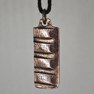 May include: Bronze rectangular pendant with horizontal ridges, suspended from a black cord. The pendant has a textured surface and a loop at the top for the cord. The overall design is rustic and minimalist.